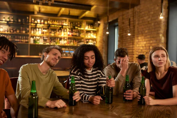 Friends looking disappointed while watching sports match on TV together, drinking beer and cheering for team in the bar. People, leisure, friendship and entertainment concept