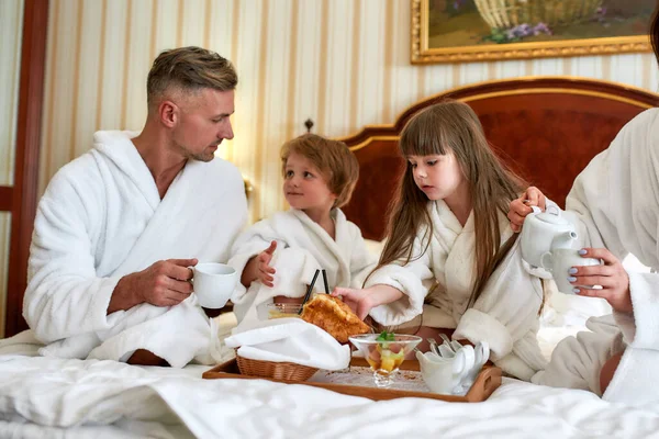 Experimente mais juntos. Pais e duas crianças em roupões de banho brancos tomando café da manhã na cama, comendo doces e bebendo café no luxuoso quarto de hotel. Família, resort, conceito de serviço de quarto — Fotografia de Stock