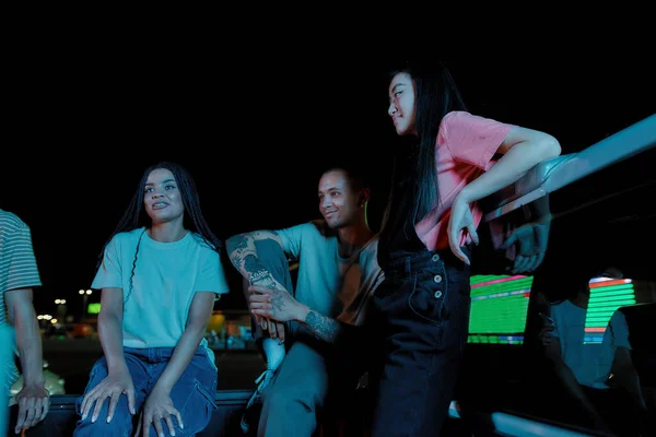 Una reunión nocturna de un grupo de jóvenes amigos bien vestidos de diferentes nacionalidades sentados en un baúl de pick-ups frente al otro afuera en un sitio de estacionamiento hablando — Foto de Stock