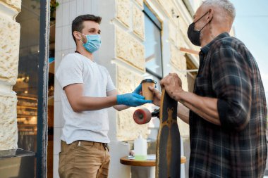 Maske takan orta yaşlı bir adam, Coronavirus tecridi sırasında yemek siparişini alırken koruyucu eldivenlerle kafe asistanının elinden sıcak kahve içiyordu.