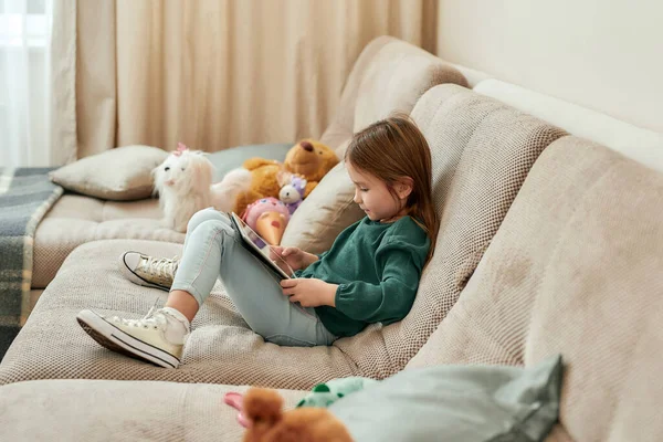 Una niña linda sentada en un sofá con sus zapatos en la celebración de una tableta —  Fotos de Stock