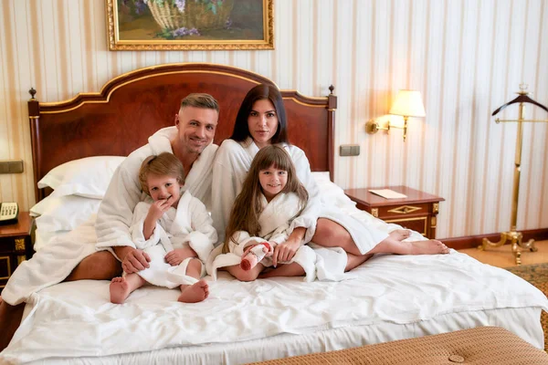 Sejam bem-vindos. Sê feliz. Pais caucasianos e duas crianças de roupão branco olhando para a câmera enquanto se sentam na cama em um luxuoso quarto de hotel. Família, viagens, resort, conceito de férias — Fotografia de Stock