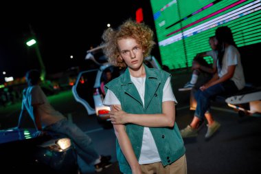 İyi giyimli, kızıl saçlı, çilli genç bir kadın. Dışarıda kameraya bakıyor. Sol eli göğsünün üstünde.