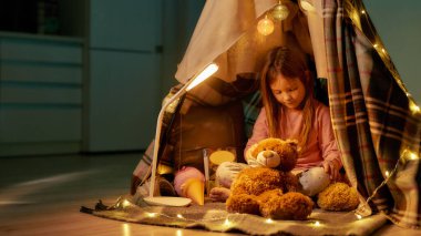 Pijama giyen küçük bir kız kendi kendini yetiştirmiş bir kulübede çıplak ayakla yerde oturuyor oyuncak ayısını soyuyordu kulübenin etrafında rahat çelenkler vardı.