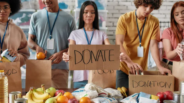 Foto recortada de un grupo de jóvenes voluntarios empacando alimentos y bebidas donación, Mujer con tarjeta de presentación de donaciones, frutas, latas de lata y paquetes sobre la mesa — Foto de Stock