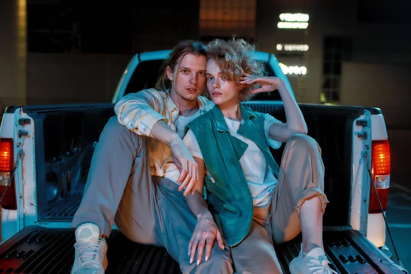 Dva mladí dobře oblečení bílí přátelé různých pohlaví hledící do kamery pózující a sedící v otevřeném kufru auta venku na parkovišti — Stock fotografie