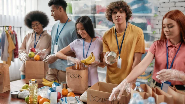 Joven voluntario mirando la cámara mientras empaca la donación de alimentos y bebidas en bolsas de papel y caja para personas necesitadas, equipo pequeño que trabaja en la fundación benéfica — Foto de Stock