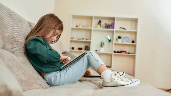 Uma menina bonita jogando videogames em um tablet sentado em um sofá — Fotografia de Stock