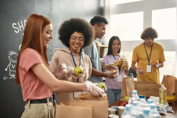 Glada unga volontärer i handskar samla in, sortera mat för behövande människor, Diverse team arbetar tillsammans med donation projekt i välgörenhet organisationskontor — Stockfoto