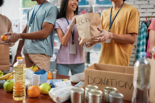 Foto recortada de voluntarios masculinos y femeninos sosteniendo bolsa de papel con comida mientras empacan la donación para personas necesitadas, equipo joven que trabaja en la fundación benéfica — Foto de Stock