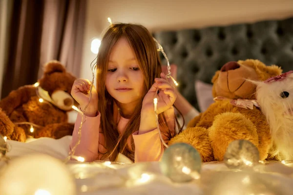 Malá roztomilá dívka leží sama na břiše na velké posteli bosá s její věnce na hlavě a v rukou s medvídky kolem — Stock fotografie