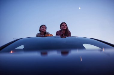 Film zamanı. Akşamları açık hava sinemasında film izlerken elinde patlamış mısır ve içeceklerle arabanın çatısında uzanan iki kız.