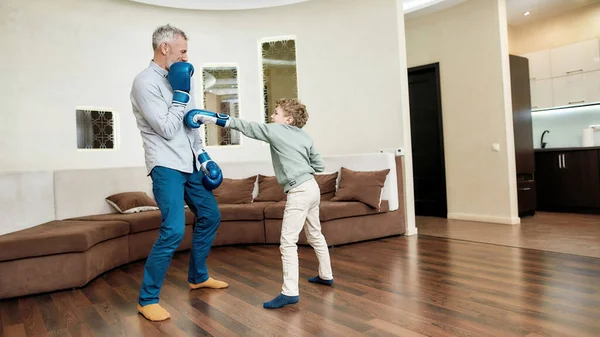 Een gezonde levensstijl. Grootvader die bokshandschoenen draagt terwijl hij bokst met zijn kleinzoon in de woonkamer — Stockfoto
