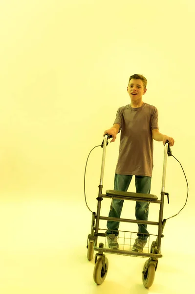 Full length shot of cheerful teenaged disabled boy with cerebral palsy looking at camera, taking steps with his walker isolated over yellow light background — Stock Photo, Image