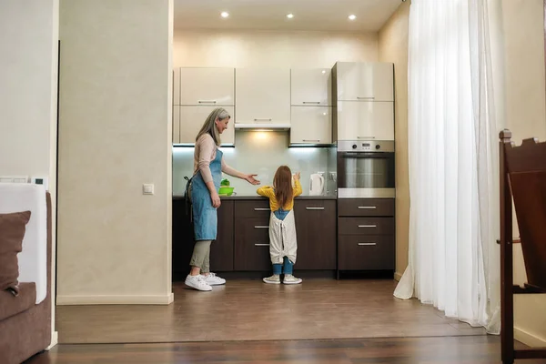 Grandmother cooking with caucasian granddaughter wearing kitchen apron in modern kitchen at home — Stock Photo, Image