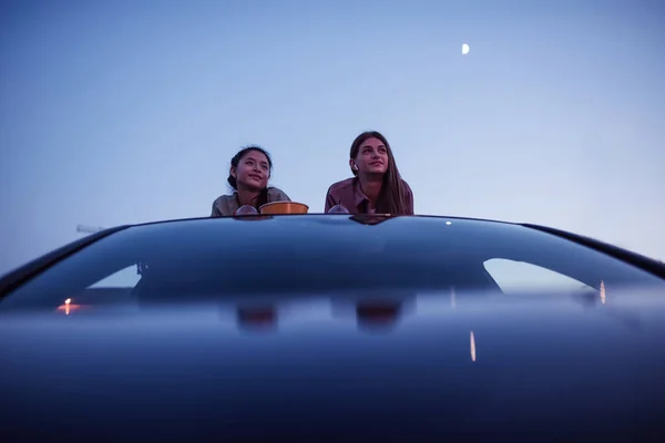 Its movie time. Two girls lying on the roof of a car with popcorn bags and drinks while watching a movie in an open air cinema in the evening