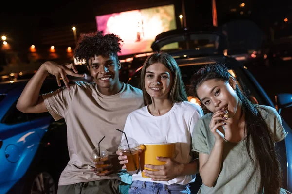 Retrato de tres alegres jóvenes amigos sonriendo a la cámara mientras están de pie juntos junto al coche estacionado frente a una pantalla grande para ver películas — Foto de Stock