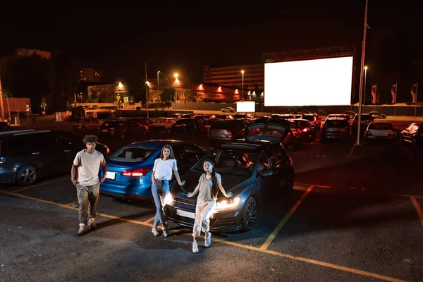 Full length shot of three diverse young friends looking at camera while standing by the car parked in front of a big screen, ready to watch a movie at drive in cinema — Stock Photo, Image