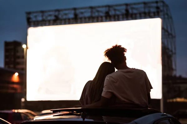 Silueta pohled na atraktivní mladý pár, chlapec a dívka objímající, trávit čas spolu, sedí v autě při sledování filmu v jízdě v kině — Stock fotografie