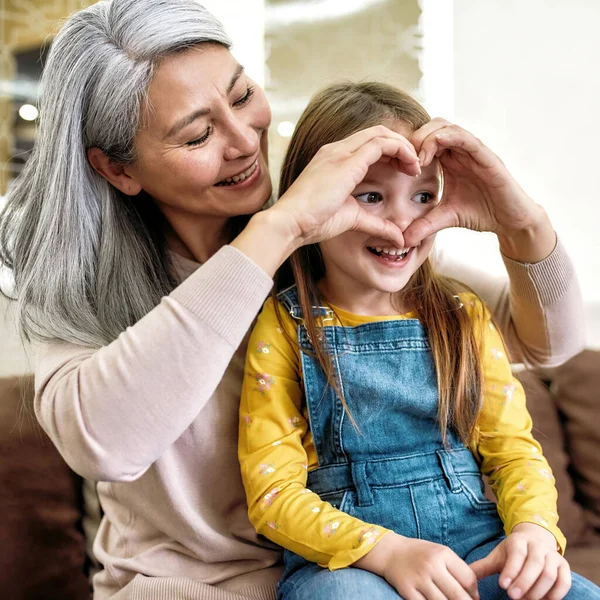 Granny loves you. Close up portrait of happy loving grandmother making heart sign while playing together with her cute little granddaughter at home