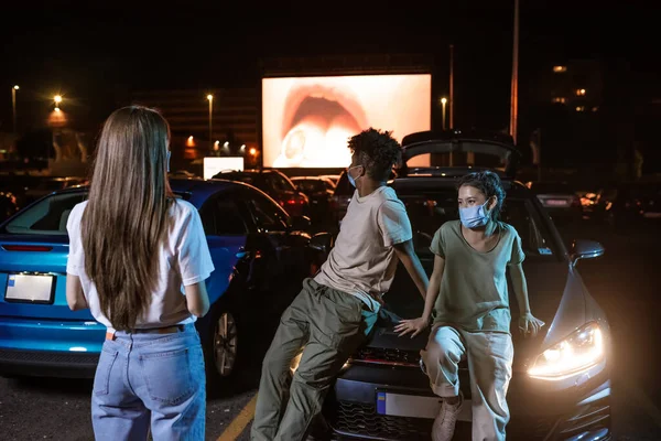 Tři různí přátelé v ochranných maskách, stojící u auta zaparkovaného před velkou obrazovkou. Mladí kluci sledují film v kině pod širým nebem — Stock fotografie