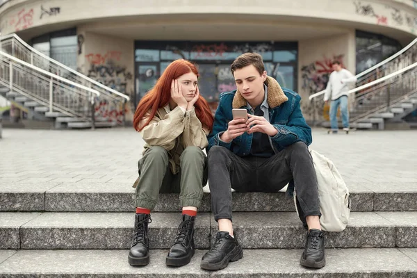 Δύο έφηβοι περνούν χρόνο μαζί, κάθονται στα σκαλοπάτια έξω. Αναστατωμένη κοπέλα κοιτάζει το αγόρι της ενώ αυτός την αγνοεί, χρησιμοποιώντας το smartphone του — Φωτογραφία Αρχείου