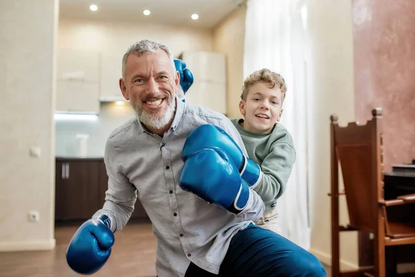 Ritratto di un nonno allegro e di un nipotino che indossano guanti da boxe blu divertendosi e sorridendo alla macchina fotografica mentre fanno allenamento di boxe — Foto Stock