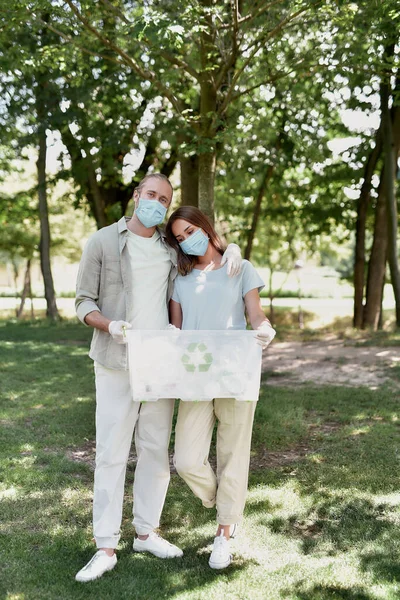 Junges Paar mit Mundschutz hält Papierkorb in der Hand und umarmt sich, sammelt Plastikmüll im Wald wegen COVID19-Pandemie — Stockfoto