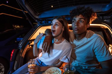Güzel genç çift film izlerken duygusal görünüyor, açık hava sinemasında büyük bir ekranın önünde arabanın bagajında birlikte oturuyorlar.