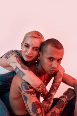 Çok kültürlü bir ilişki. Sırtında iki dövmesi olan koyu tenli üstsüz bir erkeğe sarılan genç, gülümseyen, piercing takmış bir kadın.