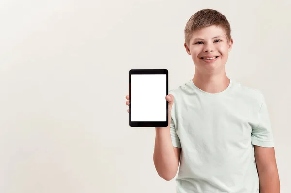 Щасливий хлопчик з синдромом Дауна посміхається, тримає і показує планшетний ПК з порожнім екраном, стоячи ізольовано на білому тлі — стокове фото