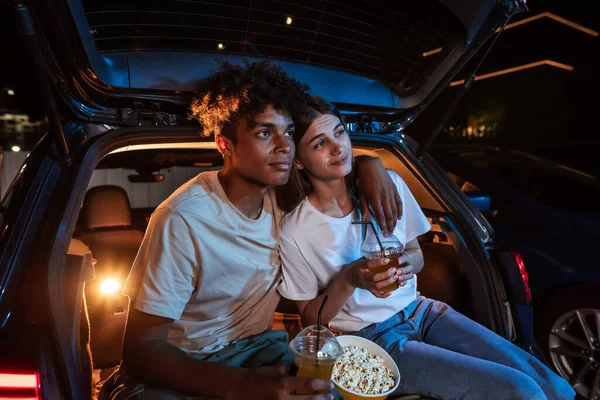 Piękna młoda para obejmująca się, oglądająca film, jedząca popcorn siedząc razem w bagażniku samochodu przed dużym ekranem w kinie na świeżym powietrzu — Zdjęcie stockowe