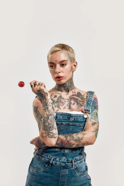 Tatuagem e piercing. Uma mulher branca perfurada com tatuagens usando uma ganga geral segurando um chupa-chupa e olhando para o lado — Fotografia de Stock