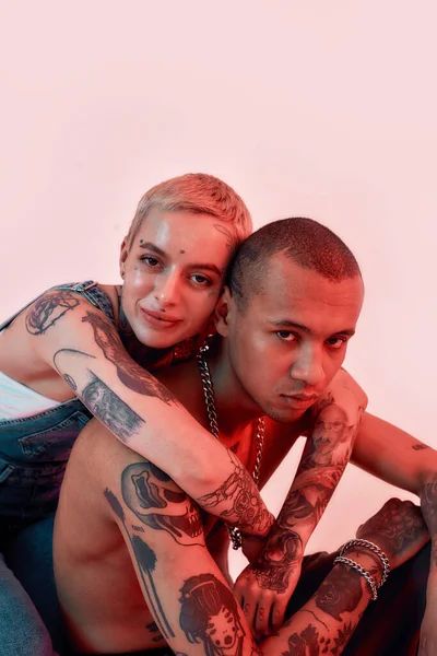 Çok kültürlü bir ilişki. Sırtında iki dövmesi olan koyu tenli üstsüz bir erkeğe sarılan genç, gülümseyen, piercing takmış bir kadın.