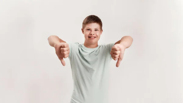 Alegre niño discapacitado con síndrome de Down sonriendo a la cámara, mostrando los pulgares hacia abajo con dos manos mientras está de pie aislado sobre fondo blanco — Foto de Stock