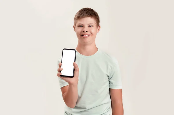 Niño discapacitado feliz con síndrome de Down sonriendo, sosteniendo y mostrando el teléfono inteligente con la pantalla en blanco en la cámara mientras está de pie aislado sobre fondo blanco — Foto de Stock