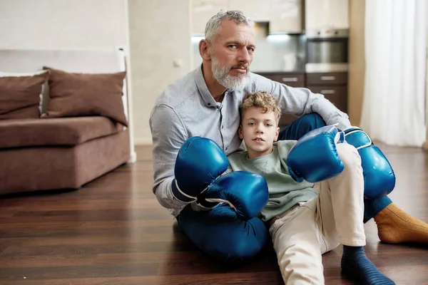 Ontspannen na de training thuis. Grootvader en kleinzoon die bokshandschoenen dragen op de vloer en naar de camera kijken — Stockfoto