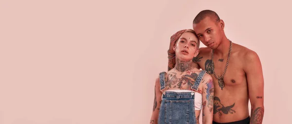 Ragazzo in topless dalla pelle scura con tatuaggi in piedi dietro una ragazza adulta tatuata bianca trafitta, abbracciandola da un retro, guardando in una fotocamera davanti a uno sfondo rosa — Foto Stock