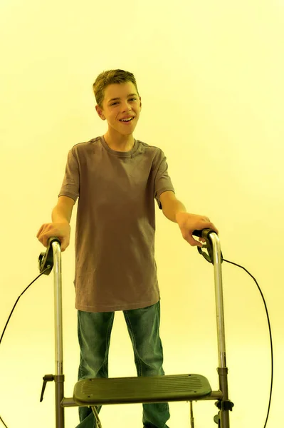 Portrait d'un adolescent handicapé joyeux avec une paralysie cérébrale souriant à la caméra, prenant des mesures avec son marcheur isolé sur fond de lumière jaune — Photo