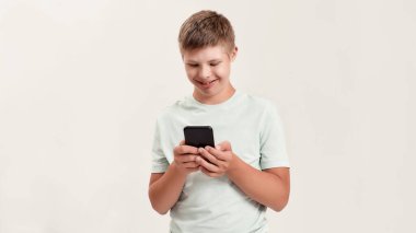 Down sendromlu, akıllı telefon kullanan genç engelli bir çocuk beyaz arka planda tek başına dikilirken ekrana bakıyor.