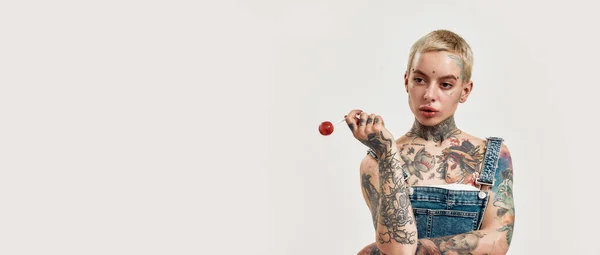 Tatuagem e piercing. Uma mulher branca perfurada com tatuagens vestindo uma ganga geral segurando um pirulito vermelho enquanto olha para os lados — Fotografia de Stock