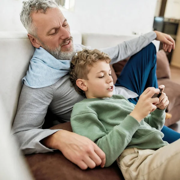 子供やガジェット。かわいい男の子は彼の幸せな祖父とソファの上でリラックスしながらスマートフォンでビデオゲームをプレイ — ストック写真