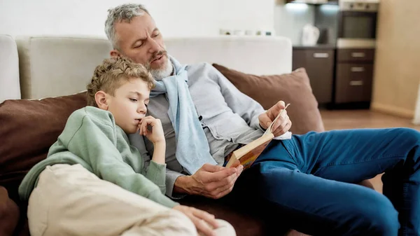 Делать домашнее задание вместе. Дедушка и внук сидят на диване в гостиной и читают вместе книгу. — стоковое фото