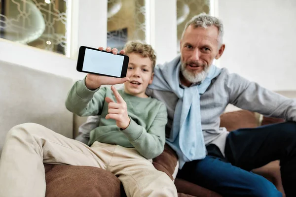 Söt liten pojke som pekar på tom mobiltelefon skärm, medan koppla av på soffan med sin farfar hemma — Stockfoto