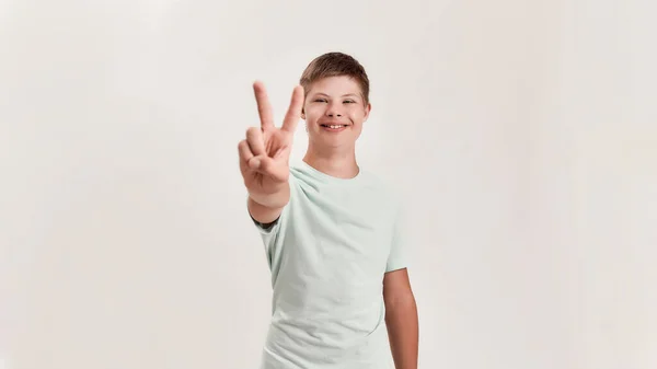 Happy disabled boy with Down syndrome smiling at camera, showing peace sign with one hand while standing isolated over white background — Stock Photo, Image