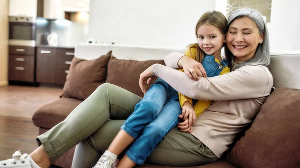 Щасливі миті. Портрет веселої бабусі і чарівної онуки, яка посміхається на камеру, обіймаючись, сидячи разом на дивані вдома — стокове фото