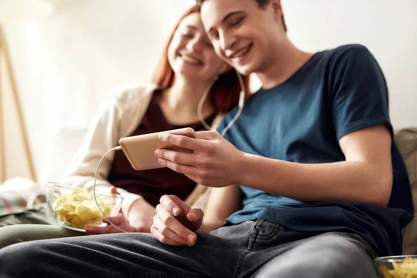Un par de adolescentes felices sentados en el sofá en casa, viendo algo por teléfono, usando el mismo par de auriculares mientras pasan tiempo juntos —  Fotos de Stock