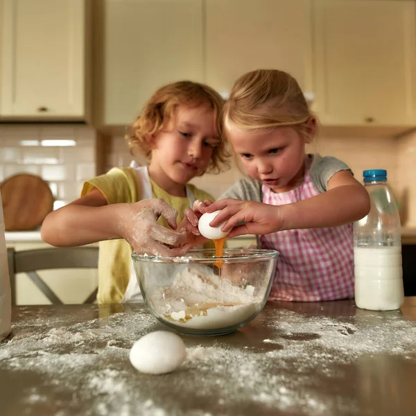 Yemek zamanı. İki küçük tatlı çocuk, önlüklü kız ve erkek yumurta eklerken odaklanıyorlar, evdeki mutfak masasında hamuru birlikte hazırlıyorlar. — Stok fotoğraf