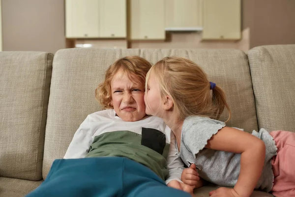 Portrét malého chlapce, který vypadá nespokojeně, zatímco jeho sourozenecká sestra ho líbá na tvář, tulí se k sobě na pohovce doma — Stock fotografie