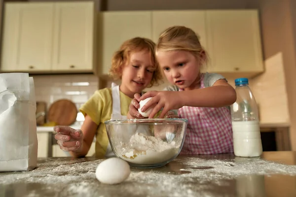 İki küçük tatlı çocuk, önlüklü kız ve erkek yumurta eklerken odaklanıyorlar, evdeki mutfak masasında hamuru birlikte hazırlıyorlar. — Stok fotoğraf